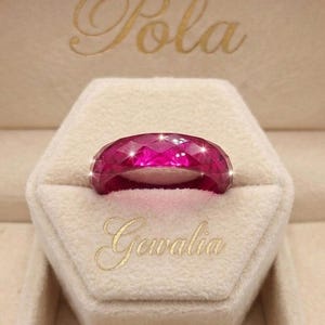 May include: A faceted, translucent magenta ring with a diamond-cut surface texture. The ring is displayed in a beige hexagonal box featuring the gold embossed text Pola and Gewalia, which is visible above and below the ring respectively.