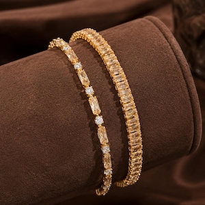 Può includere: Due bracciali color oro con pietre rettangolari taglio baguette. Un bracciale presenta una fila continua di pietre baguette, mentre l'altro alterna singole pietre taglio baguette a piccole pietre rotonde dal taglio brillante.