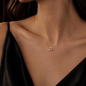 May include: A delicate gold chain necklace featuring a pendant composed of two small, interlocking circular rings. Each ring is encrusted with tiny, clear, sparkling stones that catch the light, creating a refined and subtle metallic accessory.