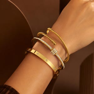 Könnte beinhalten: Ein Set aus drei goldfarbenen Armreifen. Der oberste Armreif hat eine gedrehte Kordelstruktur. Der mittlere Armreif ist ein schmales Band mit einer Reihe kleiner, klarer Steine. Der untere Armreif ist flach und mit kleinen Blumenmotiven besetzt.