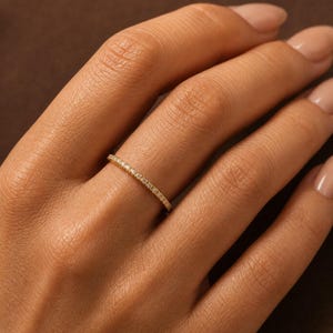 May include: A thin gold band ring featuring a continuous row of small, clear, round-cut stones set along the outer surface. The metal has a polished finish, and the stones are evenly spaced, creating a delicate and uniform appearance.