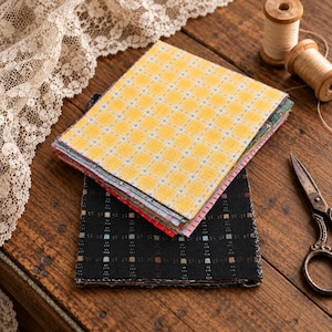 May include: A stack of square fabric swatches featuring a yellow plaid pattern on top and a black grid pattern on the bottom, accompanied by two wooden thread spools, a pair of metal scissors, and a piece of white lace trim.
