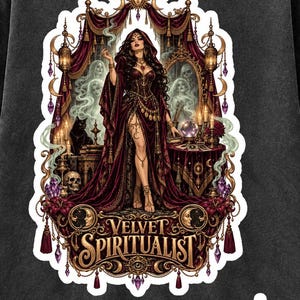 Puede incluir: Una camiseta gris carbón con un gráfico detallado de una figura con túnica roja oscura frente a un espejo y una mesa con una bola de cristal. El diseño incluye un gato negro, velas y el texto VELVET SPIRITUALIST en letras de color dorado.
