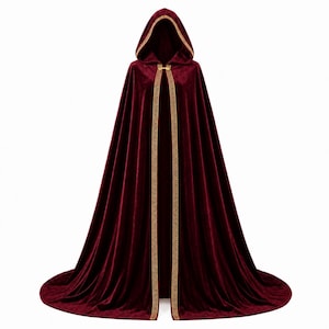 May include: A floor-length velvet cloak in a deep burgundy red color, featuring a large hood and a gold-colored metallic clasp at the neck. The front edges and the hood are trimmed with a decorative gold Greek key pattern border.