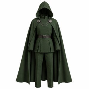 Può includere: Un mantello con cappuccio verde scuro indossato sopra una giacca abbinata a maniche lunghe con vita plissettata e pantaloni a gamba dritta. Il mantello presenta due fermagli metallici circolari color argento collegati da una doppia catena. È indossata una cintura marrone scuro.