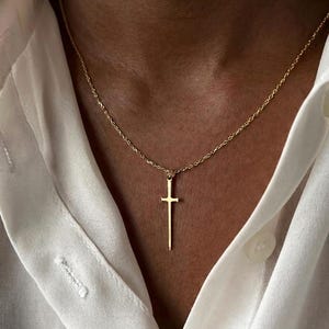 Può includere: Una collana color oro con un ciondolo sottile a forma di spada sospeso a una delicata catenina a maglie. Il ciondolo presenta una punta affilata, una guardia orizzontale e un piccolo anello nella parte superiore fissato alla catena, indossato su una camicia bianca.