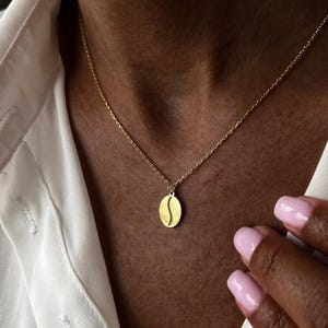 May include: A gold-toned necklace featuring a small, oval-shaped pendant with a central vertical curved cutout, resembling a coffee bean. The pendant hangs from a delicate, thin gold-toned chain that rests against the collarbone area.