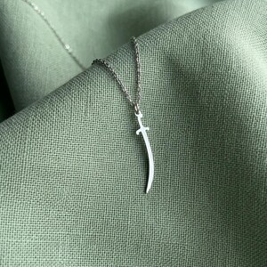 Può includere: Una collana color argento con un ciondolo a forma di spada curva o scimitarra. Il ciondolo è appeso a una sottile e delicata catenina di metallo. Il design è semplice ed essenziale, con una finitura lucida che riflette la luce.
