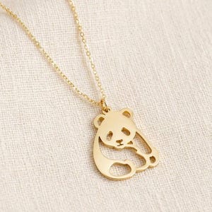 Op de afbeelding: Een goudkleurige ketting met een fijne schakelketting en een platte, uitgesneden hanger in de vorm van een zittende panda. Het panda-ontwerp is uitgevoerd in een eenvoudige, strakke omtrek met uitsparingen voor de ogen, oren en ledematen.