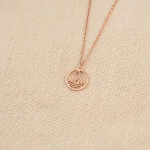 Puede incluir: Un collar de tono oro rosa con una cadena delicada y un colgante circular. El colgante presenta un diseño recortado de una forma estilizada de llama o gota de agua centrada dentro del marco redondo, suspendido de una cadena de metal fina.