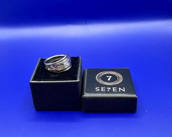May include: A silver-toned metal ring with an engraved floral pattern resting on a black velvet-lined box. Next to it is a black square box with the number 7 inside a circular design and the text SE7EN printed in white on the top surface.