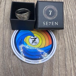 Pode incluir: Um anel de metal prateado com um padr&atilde;o floral em relevo dentro de uma caixa quadrada preta com a etiqueta SE7EN. Abaixo da caixa h&aacute; um adesivo circular com um gr&aacute;fico de onda colorido e o texto 7 Rings no centro do design.
