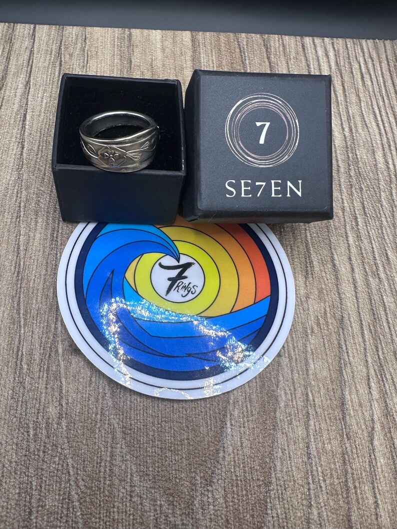 Pode incluir: Um anel de metal prateado com um padr&atilde;o floral em relevo dentro de uma caixa quadrada preta com a etiqueta SE7EN. Abaixo da caixa h&aacute; um adesivo circular com um gr&aacute;fico de onda colorido e o texto 7 Rings no centro do design.