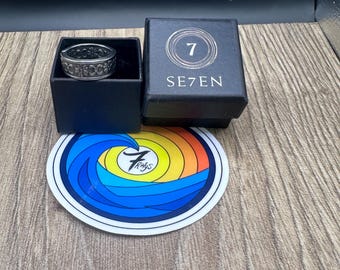 Pode incluir: Um anel de metal prateado com um padrão floral repetitivo está dentro de uma caixa quadrada preta com o número 7 e o texto SE7EN. Abaixo da caixa, há um adesivo circular com um design de onda azul e o texto 7 Rings.