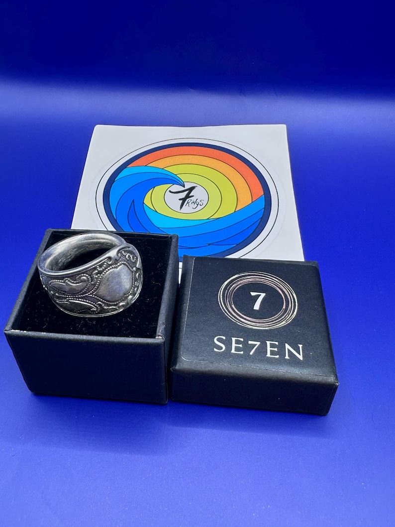 Pode incluir: Um anel de metal prateado com um padr&atilde;o floral em relevo est&aacute; numa caixa preta. Ao lado, h&aacute; uma caixa preta com a etiqueta SE7EN e o n&uacute;mero 7. Atr&aacute;s, um cart&atilde;o branco apresenta um gr&aacute;fico circular com um arco-&iacute;ris e uma onda azul.