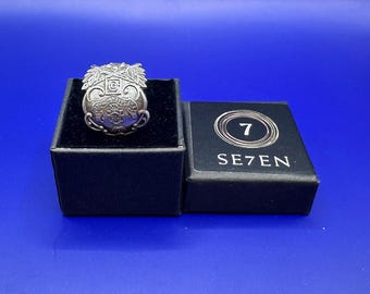 Könnte beinhalten: Ein silberfarbener Ring mit einem komplexen Wappenmotiv und Blattverzierungen, der in einer schwarz ausgekleideten Box liegt. Daneben befindet sich ein schwarzer Deckel mit der Zahl 7 in einem Kreis und dem in Weiß gedruckten Text SE7EN.