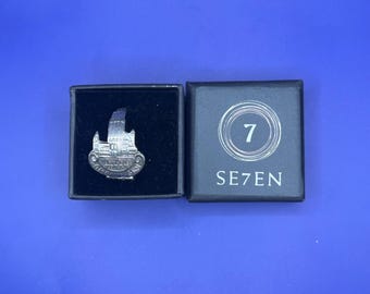 Puede incluir: Un anillo de metal plateado con un diseño arquitectónico detallado, presentado dentro de una caja cuadrada negra. La tapa de la caja está colocada al lado, mostrando un logotipo circular con el número 7 y el texto SE7EN impreso en blanco.