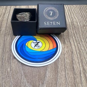 以下が含まれることがあります： エンボス加工された花柄のシルバー調の金属製リングが、SE7ENと数字の7のロゴが記された黒い正方形の箱に入っています。箱の下には、カラフルな波のデザインと黒い筆記体で7 Ringsと書かれた円形のステッカーが置かれています。
