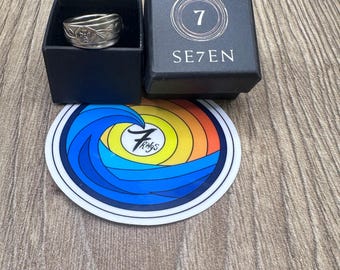Puede incluir: Un anillo de metal plateado con un patrón floral en relieve se encuentra dentro de una caja cuadrada negra etiquetada como SE7EN con un logotipo de 7. Debajo de la caja hay una pegatina circular con un diseño de ola colorido y el texto 7 Rings.