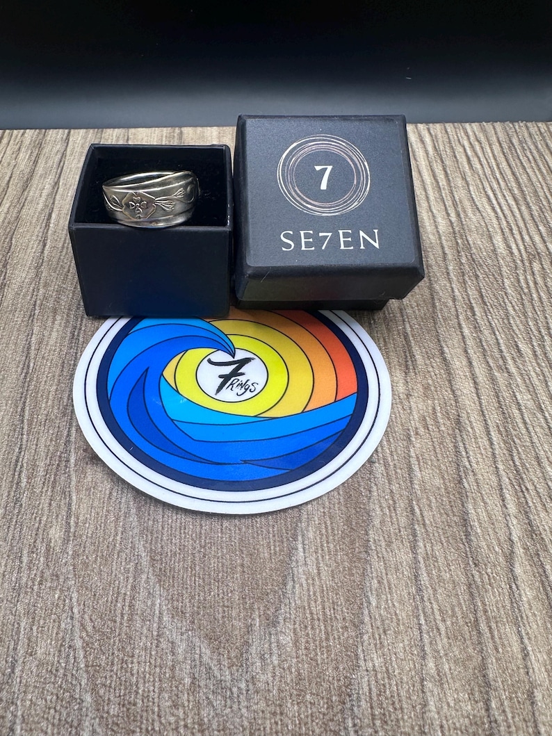 Pode incluir: Um anel de metal prateado com um padr&atilde;o floral em relevo est&aacute; dentro de uma caixa quadrada preta com a etiqueta SE7EN e um log&oacute;tipo com o n&uacute;mero 7. Abaixo da caixa est&aacute; um autocolante circular com um design de onda colorido e o texto 7 Rings.