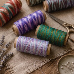 May include: Four wooden spools of variegated thread in red-green, purple, teal-gold, and lavender-blue colourways, accompanied by a pair of metal scissors and a small dish containing several straight sewing pins with white heads.