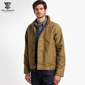May include: A tan canvas jacket with a brown corduroy collar, featuring a front zip closure, a button-down placket, and long sleeves. The jacket is worn over a blue denim button-down shirt and dark blue jeans. The logo JTC The Jackets Company is visible.