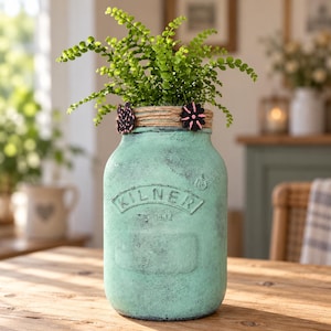 May include: A light green, distressed-finish glass jar with the embossed text KILNER on the front. The neck is wrapped in natural jute twine, decorated with two small, dark, flower-shaped wooden accents. A green fern plant grows from the jar.