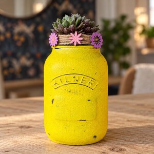 May include: A bright yellow painted glass jar with a textured finish and the embossed text KILNER. The jar is wrapped at the top with brown twine decorated with small pink and purple flower cutouts and contains a green succulent plant.