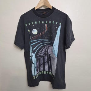 May include: A charcoal gray short-sleeve t-shirt featuring a graphic print of a bridge under a starry night sky. The text SOUNDGARDEN appears in light green at the top, and SUPERUNKNOWN is printed in light green at the bottom of the graphic.