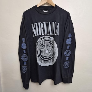 May include: A black long-sleeve t-shirt featuring the word NIRVANA in white text at the top. The centre graphic depicts a circular diagram labelled with circles of hell. Both sleeves feature light blue geometric and circular graphic prints.