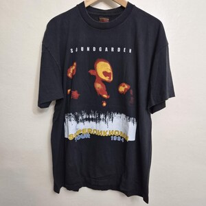 May include: A black short-sleeve t-shirt featuring the text SOUNDGARDEN at the top. The center graphic displays abstract red and yellow shapes above a white horizontal band with the text SUPERUNKNOWN TOUR 1994 printed in yellow and blue.
