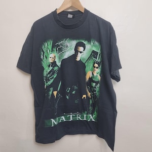 Könnte beinhalten: Ein anthrazitfarbenes Kurzarm-T-Shirt mit einem Grafikdruck von drei Charakteren aus dem Film The Matrix. Das Design zeigt grüne Lichteffekte, städtische Gebäude und den weißen Schriftzug THE MATRIX am unteren Rand der Grafik.