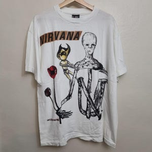 Puede incluir: Una camiseta blanca de manga corta con un estampado gráfico en la parte delantera. El diseño incluye el texto NIRVANA en letras marrones, una figura estilizada, una pequeña criatura con cuernos y una flor roja. La palabra INCESTICIDE aparece en negro.