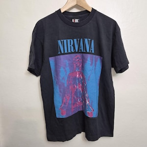 Puede incluir: Una camiseta negra de manga corta con la palabra NIRVANA en letras azul claro sobre un gráfico de una figura humana en tonos rosa y azul. La camiseta tiene cuello redondo y una etiqueta negra en el cuello con la palabra giant en color blanco.