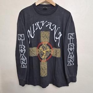 Puede incluir: Una camiseta de manga larga gris carbón con la palabra NIRVANA en texto blanco en el pecho. Un gran gráfico de cruz celta amarilla y negra con un contorno circular rojo está centrado debajo del texto. La palabra NIRVANA está impresa verticalmente en blanco en ambas mangas.