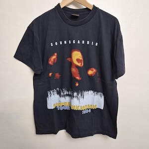 May include: A black short-sleeve t-shirt featuring the text SOUNDGARDEN at the top. The center graphic displays a stylized orange and yellow face with abstract shapes. Below, a white textured band contains the text SUPERUNKNOWN TOUR 1994.