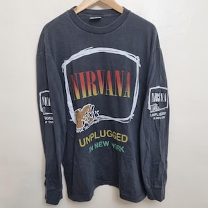 Könnte beinhalten: Ein anthrazitfarbenes Langarmshirt mit einem weißen, handgezeichneten quadratischen Rahmen um den Text NIRVANA in rot-gelben Verlaufsbuchstaben. Darunter befindet sich ein MTV-Logo und der Text UNPLUGGED IN NEW YORK in gelber und grüner Schrift.