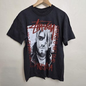 Puede incluir: Una camiseta negra de manga corta con un estampado gráfico en blanco y negro de Kurt Cobain. El diseño incluye el logotipo rojo de Stussy en la parte superior, el texto rojo "GRUNGE IS NOT DEAD" a la derecha y "NIRVANA" en rojo abajo.