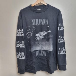 May include: A charcoal grey long-sleeve t-shirt featuring a black and white graphic of a person playing guitar on the chest. The text NIRVANA appears above the graphic, BLEW below it, and the text SUB POP is printed repeatedly down both sleeves.