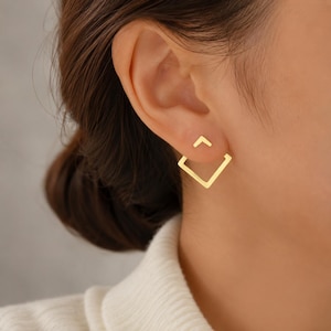 Può includere: Un orecchino ear jacket color oro caratterizzato da un piccolo perno a forma di chevron sul davanti e da un componente più grande a forma di quadrato aperto posizionato dietro il lobo, creando un effetto geometrico a strati con finitura lucida.