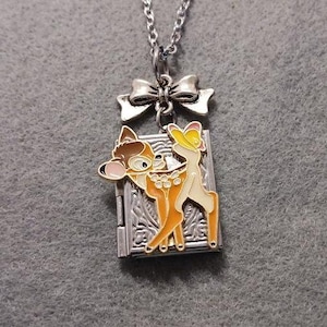 Puede incluir: Collar plateado con un colgante que presenta un diseño colorido de Bambi y Tambor. El colgante es rectangular con un patrón adornado y un lazo en la parte superior. La cadena es de estilo eslabón delicado.