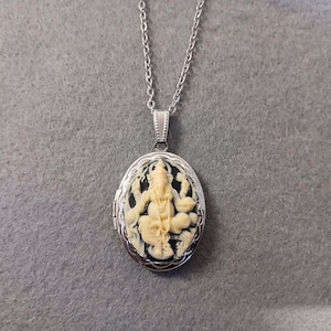 Puede incluir: Un collar de cadena de plata con un medallón ovalado que presenta un camafeo del dios hindú Ganesha. El camafeo es blanco sobre un fondo negro.