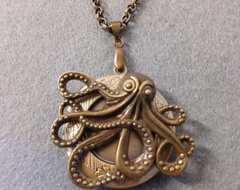 Op de afbeelding: Een bronzen octopus-hangerketting. De octopus is versierd met kleine kralen en heeft een rond medaillon in het midden. De ketting is aan een ketting.