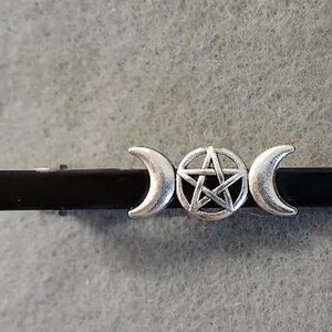 May include: A black metal bar with silver-colored crescent moons and a pentagram symbol. The bar is a tie clip, designed to hold a tie in place. The clip is a decorative accessory.