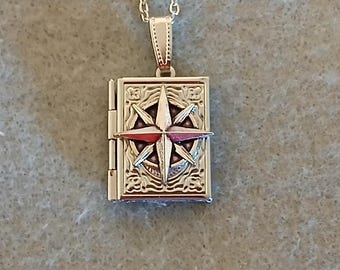 May include: A gold-toned metal pendant shaped like a small rectangular book with a hinged spine. The front cover features an embossed compass rose design with intricate scrollwork detailing. The pendant hangs from a matching gold-toned metal chain.