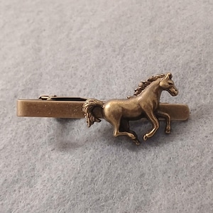 May include: A bronze tie clip with a running horse design. The horse is finely detailed, showing its mane and tail. The clip has a rectangular bar, designed to attach to a tie. The clip is approximately 5 cm in length.