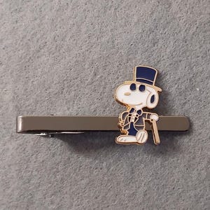 May include: A silver-coloured tie bar with a cartoon character in a top hat and suit, holding a cane. The character is white with black accents and wears a navy blue suit and hat. The tie bar is rectangular.