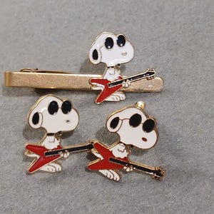 Peut inclure: Une pince à cravate dorée et deux épingles représentant Snoopy jouant d'une guitare rouge. Les figurines de Snoopy sont blanches avec des accents noirs et des lunettes de soleil. L'ensemble est sur fond gris.