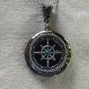 Puede incluir: Collar con colgante de brújula de plata con una piedra preciosa azul en el centro. La brújula presenta un diseño detallado con fondo negro y detalles plateados.
