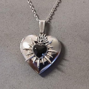 Peut inclure: Pendentif en forme de cœur argenté avec une pierre centrale noire en forme de cœur. Le pendentif présente un motif de soleil autour de la pierre noire et est suspendu à une chaîne argentée. Le médaillon est fermé.